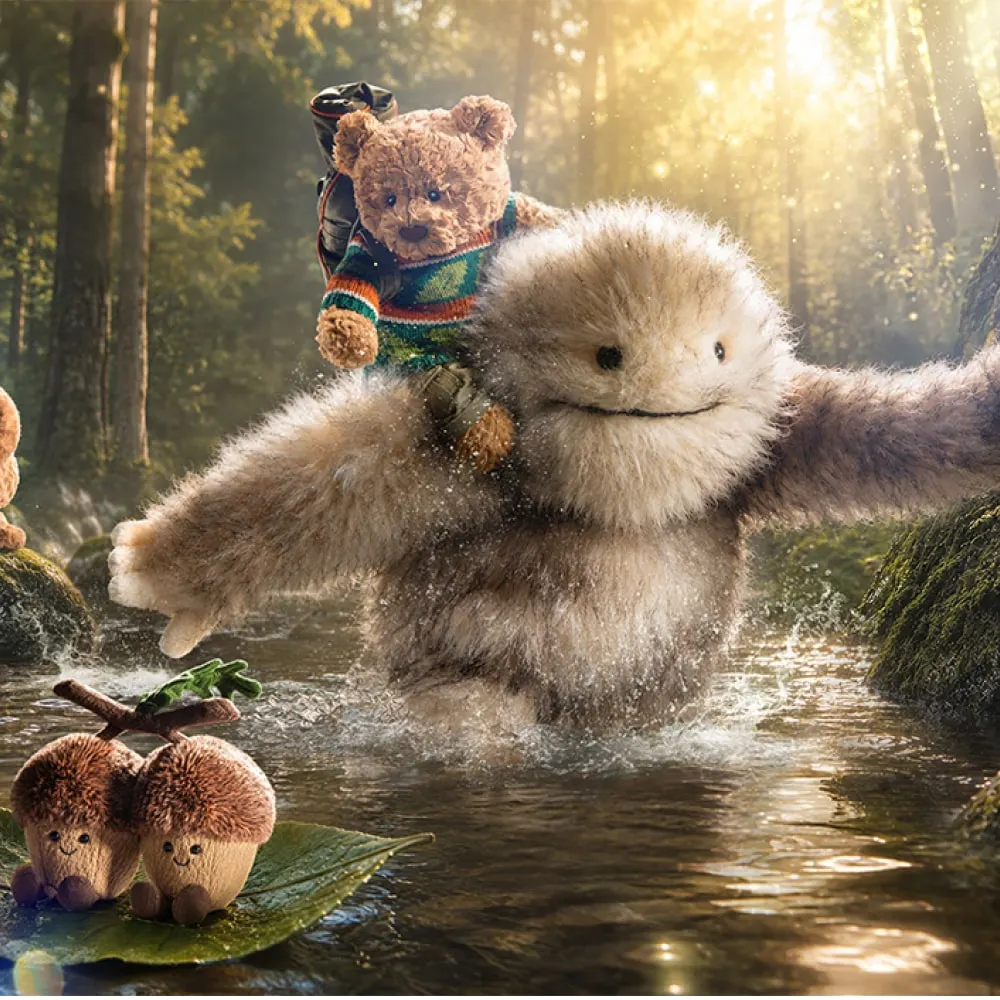 Jellycat Bartholomew Bear riding a fluffy snow giant across a woodland stream in golden light